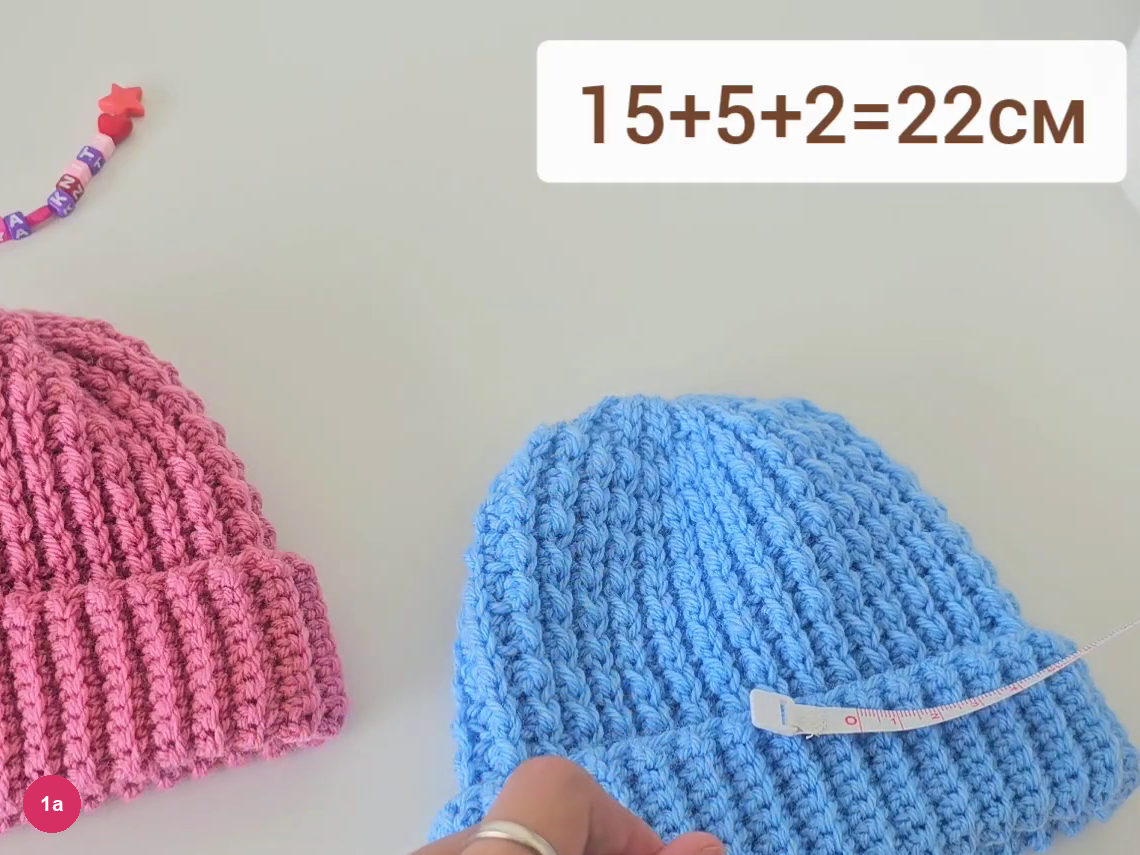 Blue ribbed hat with a tape measure laid across its folded cuff beside a pink hat, with the sum 15+5+2=22 on screen