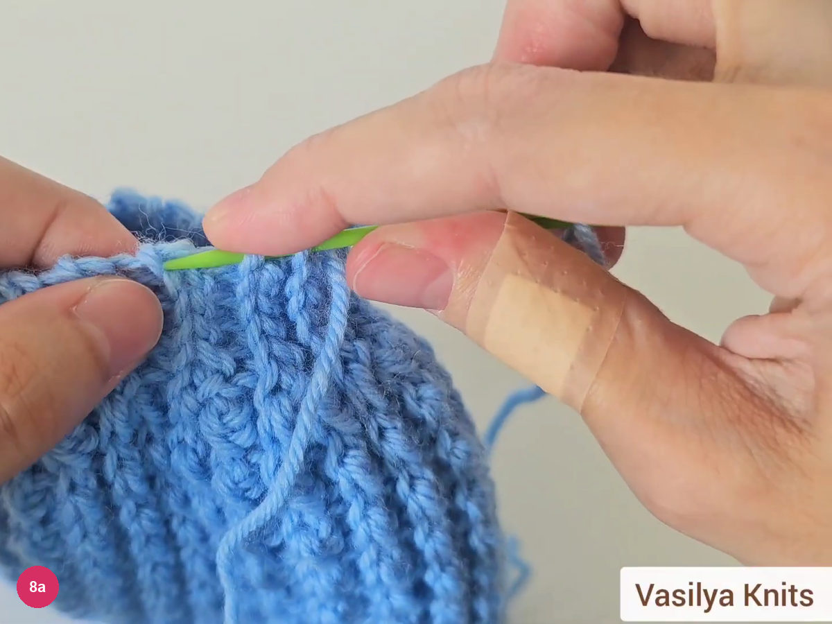 Green needle passing through the top edge loops of the blue hat with the yarn tail hanging down