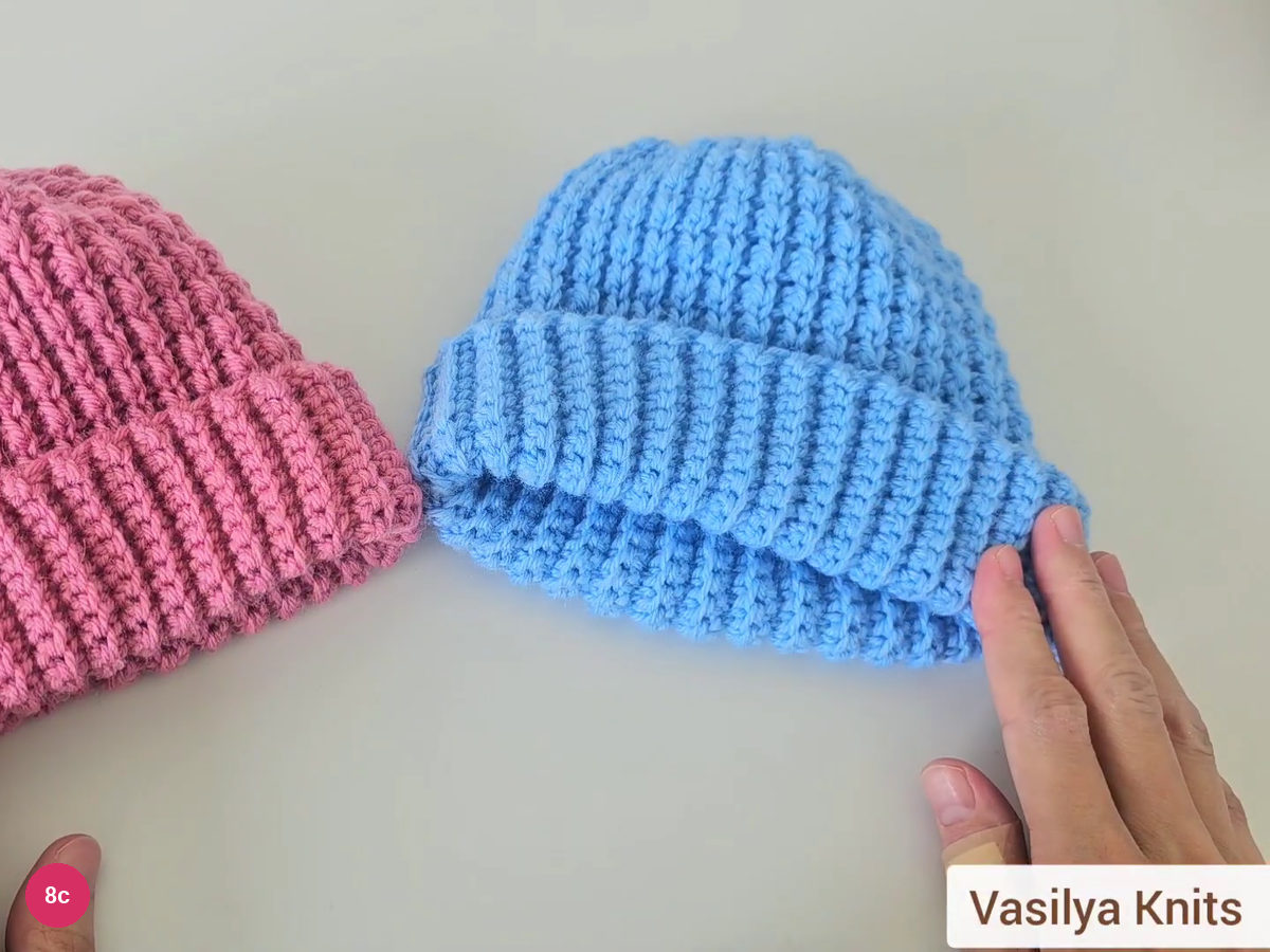 Finished blue ribbed beanie with a turned-up cuff next to a pink version, with the creator's name in the corner