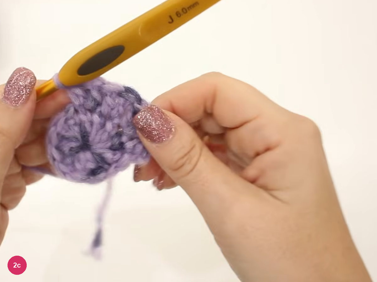 Lilac crochet disc held by fingertips with a 6.0 mm hook angled across its top edge during round two