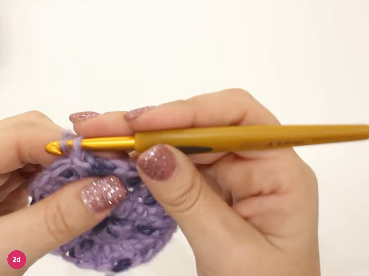 Lilac crochet disc under a horizontal gold hook while the left fingers hold the rounded edge during round three
