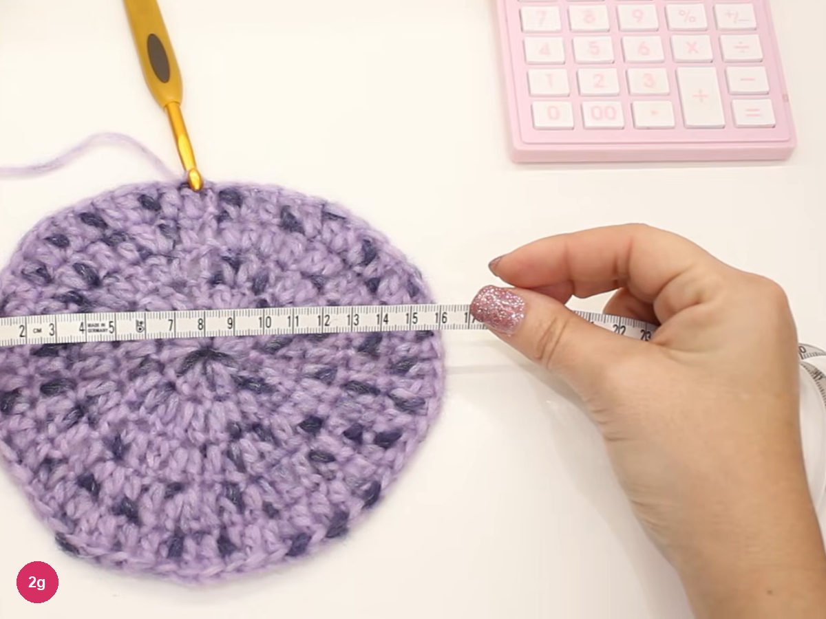 Tape measure laid across a flat lilac crochet crown, with a pink calculator and a crochet hook beside it