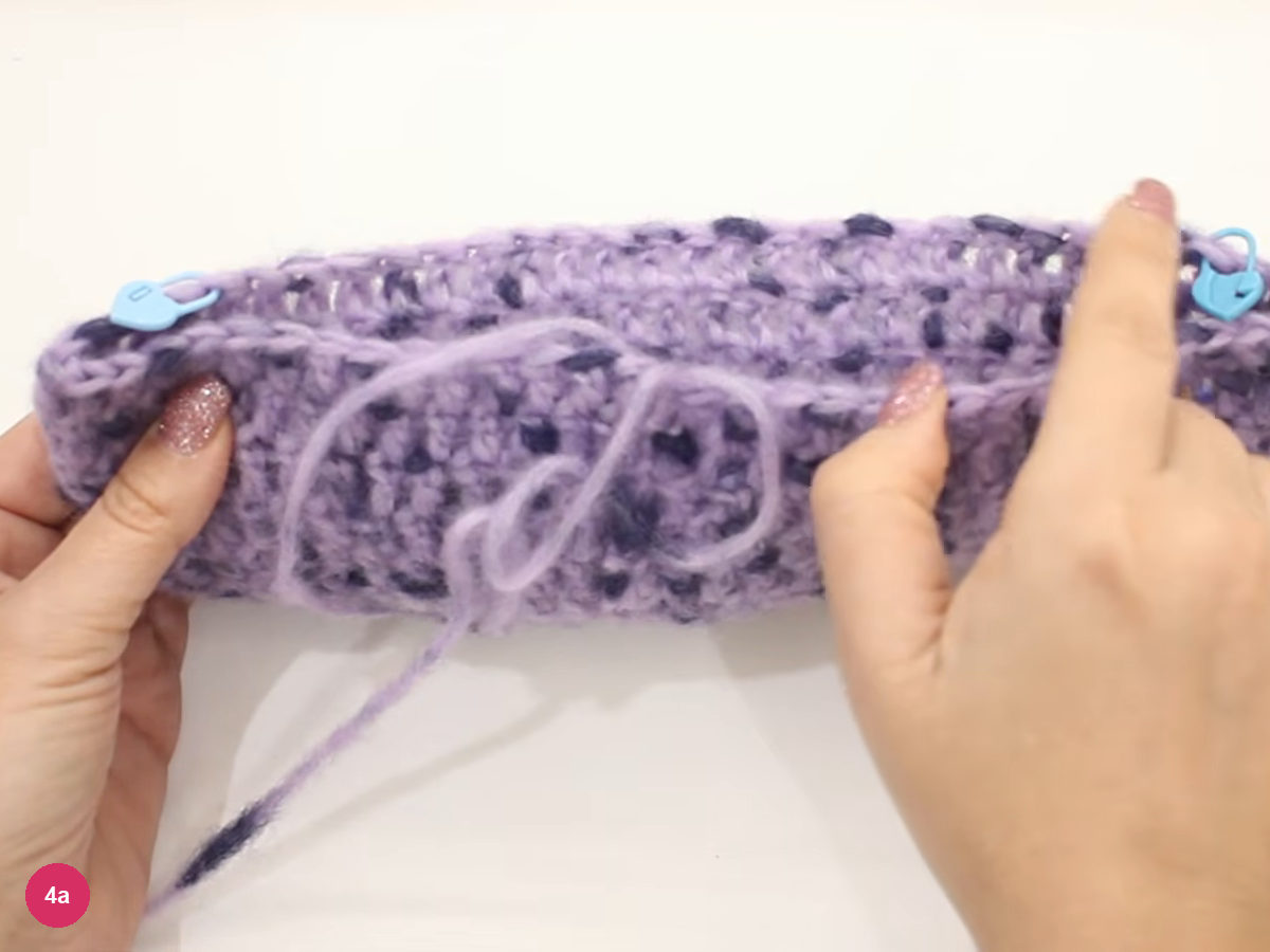 Lilac cap folded flat with a blue stitch marker at each end of its rim and a loose loop of yarn across the front