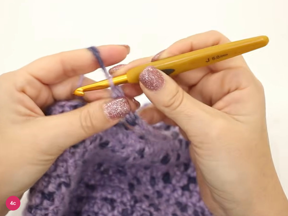 Hook and fingers drawing yarn into a chain at the rim of the lilac cap, with the cap's dotted stitches below