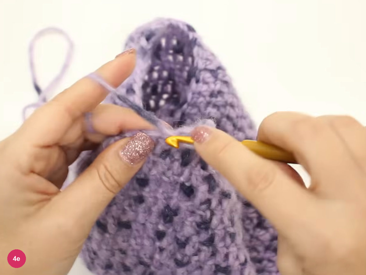 Hook entering the chain edge of the lilac hood while the yarn runs over the left forefinger