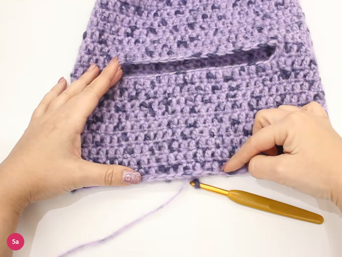 Lilac hood laid flat on a white table with a horizontal opening across it and a crochet hook beside it