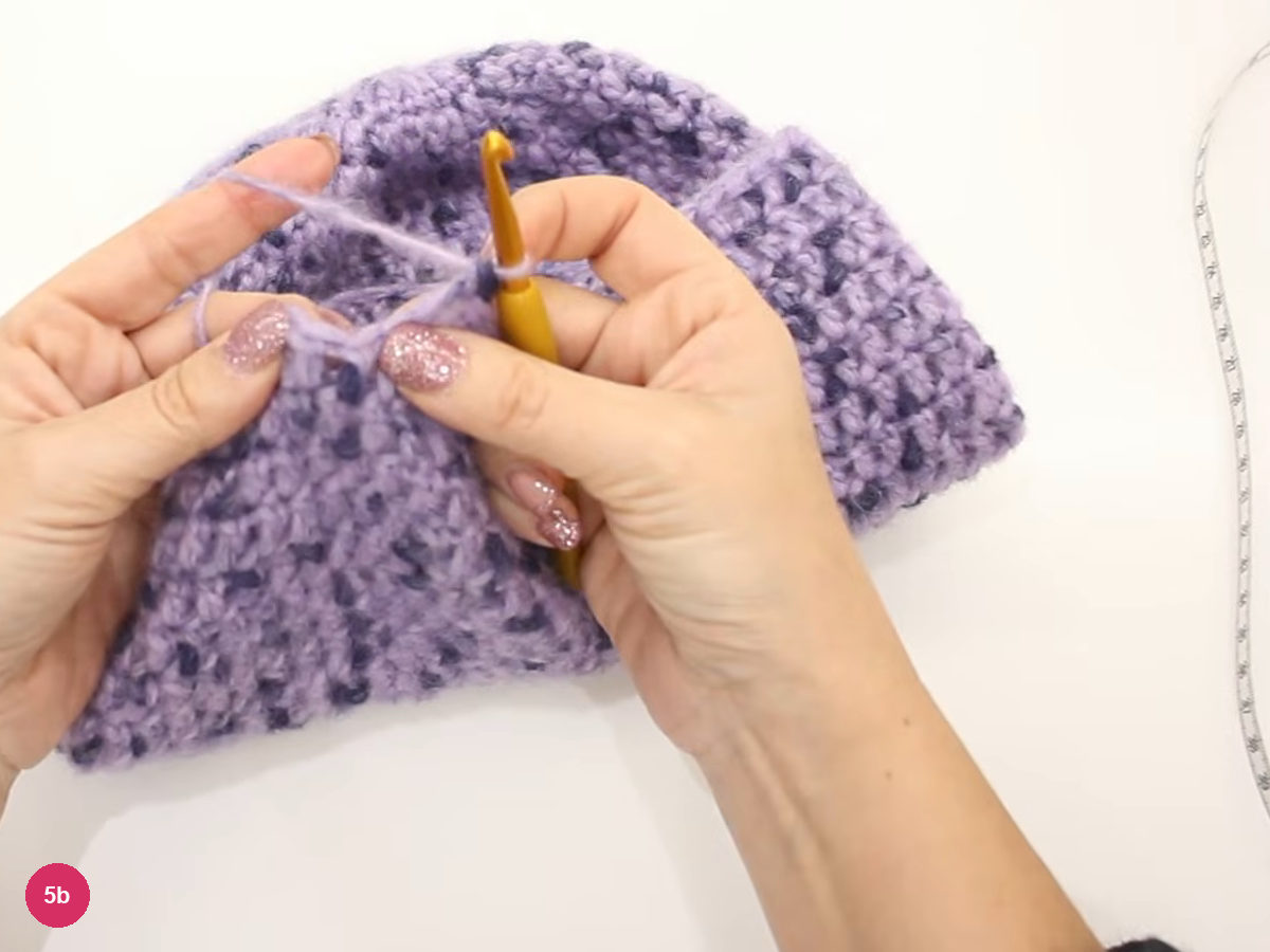 Both hands holding hook and yarn at the centre of the lilac piece, with a folded rim at upper right