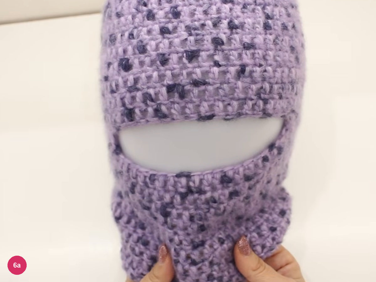 Lilac balaclava on a blank white head form seen from the front, with the opening and neck section held by two hands