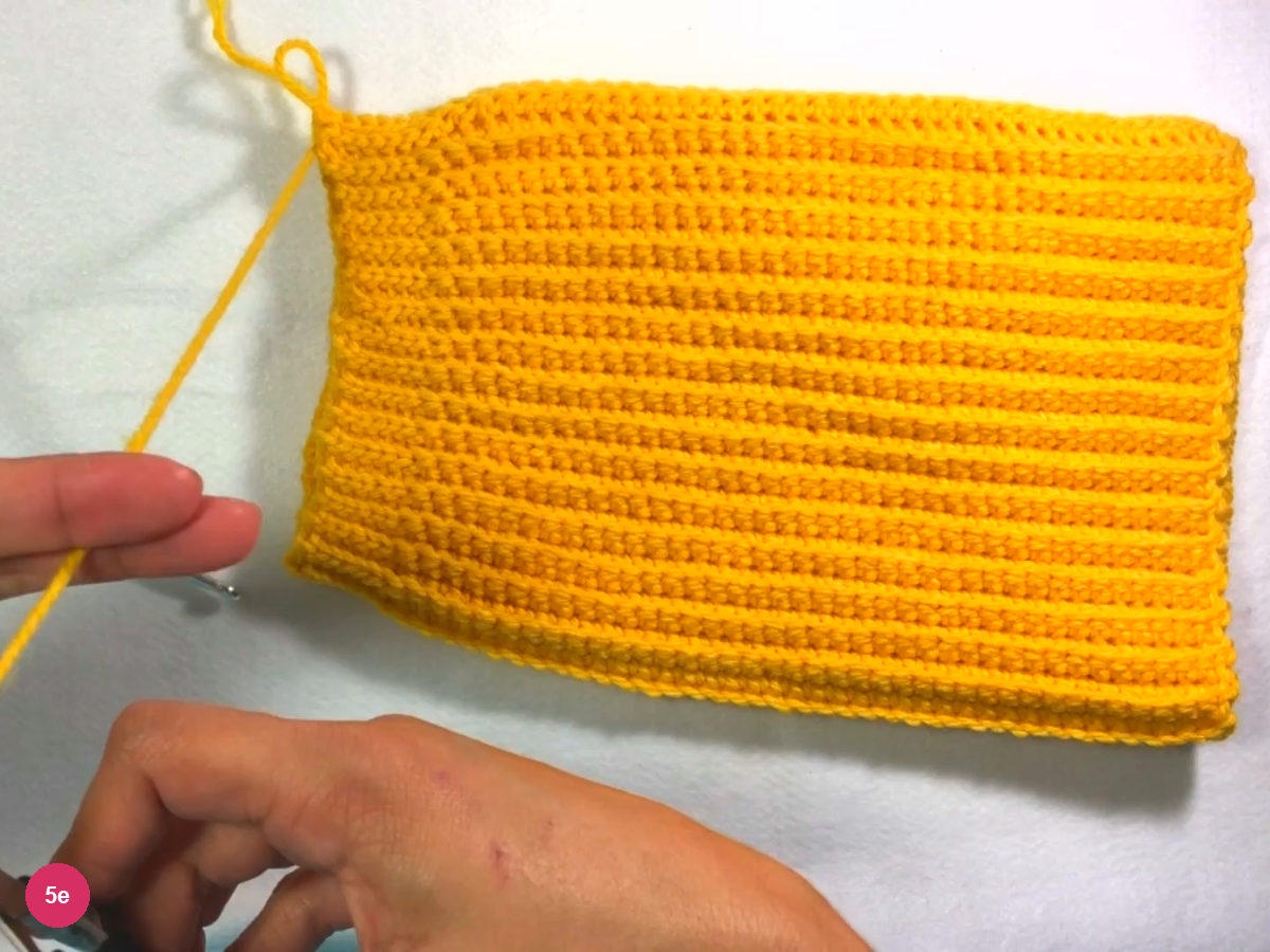 Flat ribbed yellow rectangle with the yarn tail hanging from its top left corner and a hand holding the tail