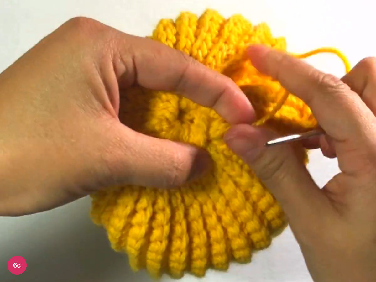 Gathered top of the beanie with ribs radiating round a small centre, the yarn trailing to the right and the needle at lower right