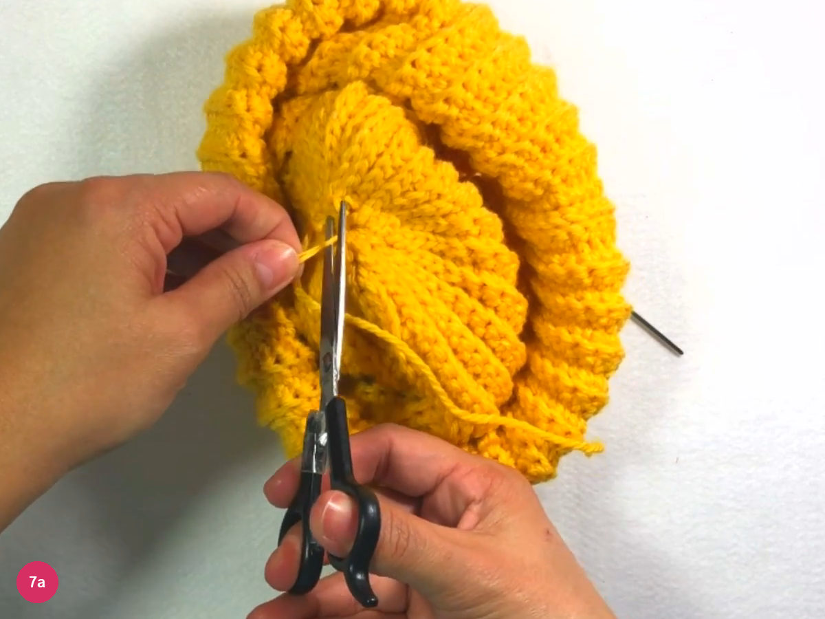 Scissors trimming the yellow yarn at the gathered crown while a hand holds the hat
