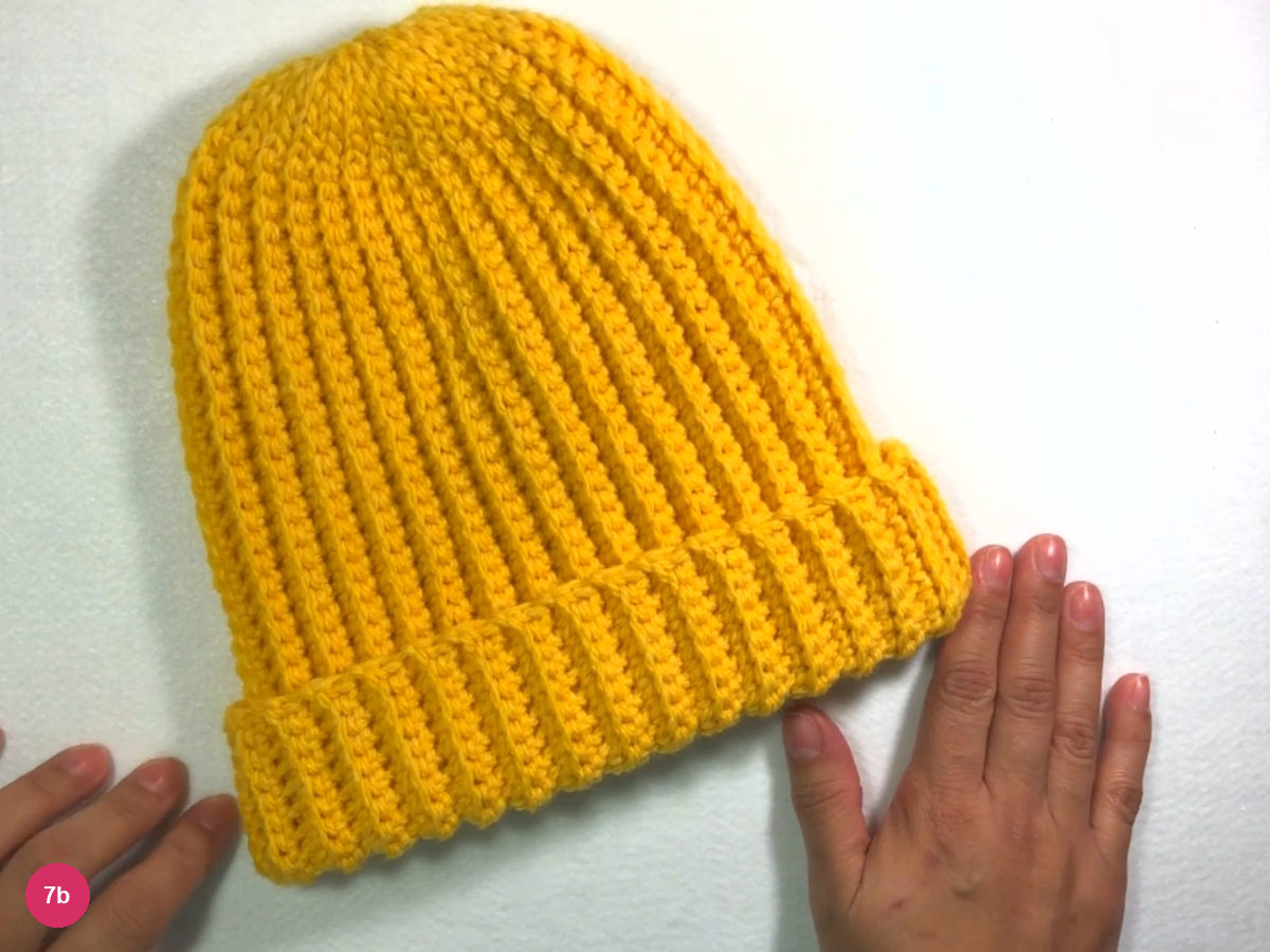 Whole finished yellow ribbed beanie laid flat on white with straight vertical ribs and a rounded crown, a hand at each side of the folded lower edge