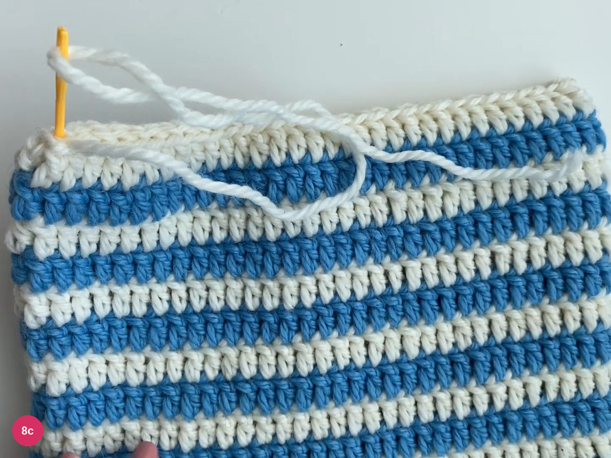Flat blue-and-white hat with both top corners, the full white seam, a yellow yarn needle and loose white seaming yarn