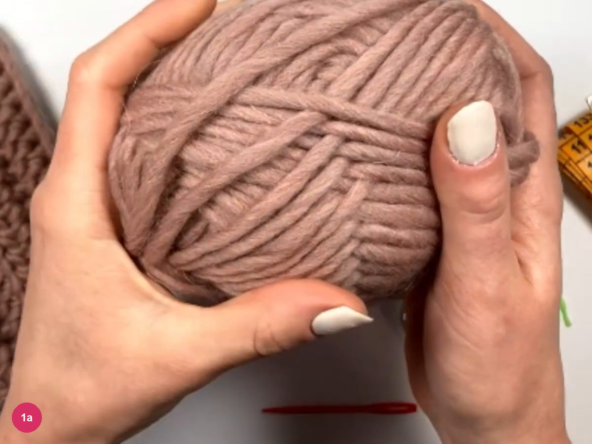 Both hands holding a ball of dusty-rose bulky wool with the on-screen yarn description above it