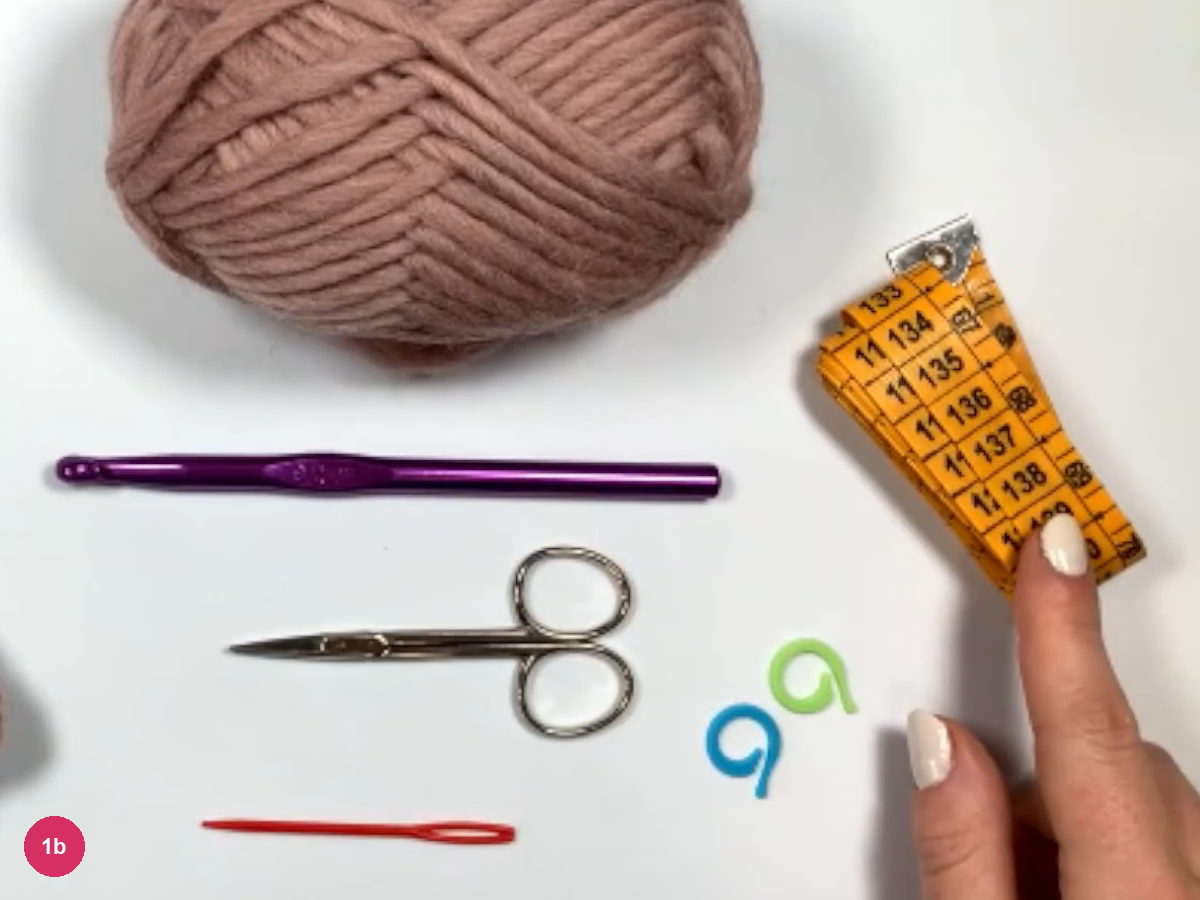 Dusty-rose yarn ball, purple crochet hook, silver scissors, two stitch markers, orange tape measure and red tapestry needle laid out on white