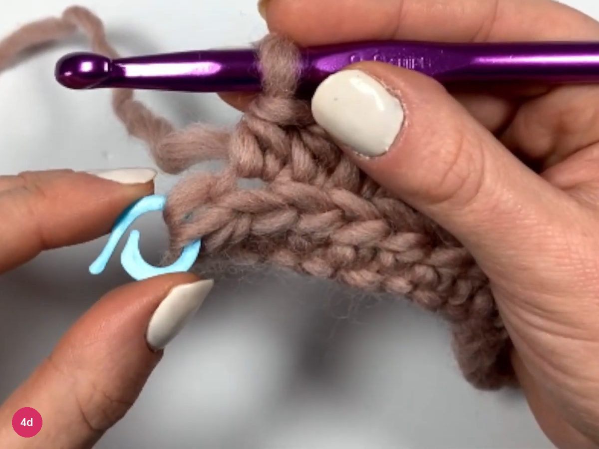 A blue stitch marker held in the fingers at the end of row 2, with the hook above the last stitch
