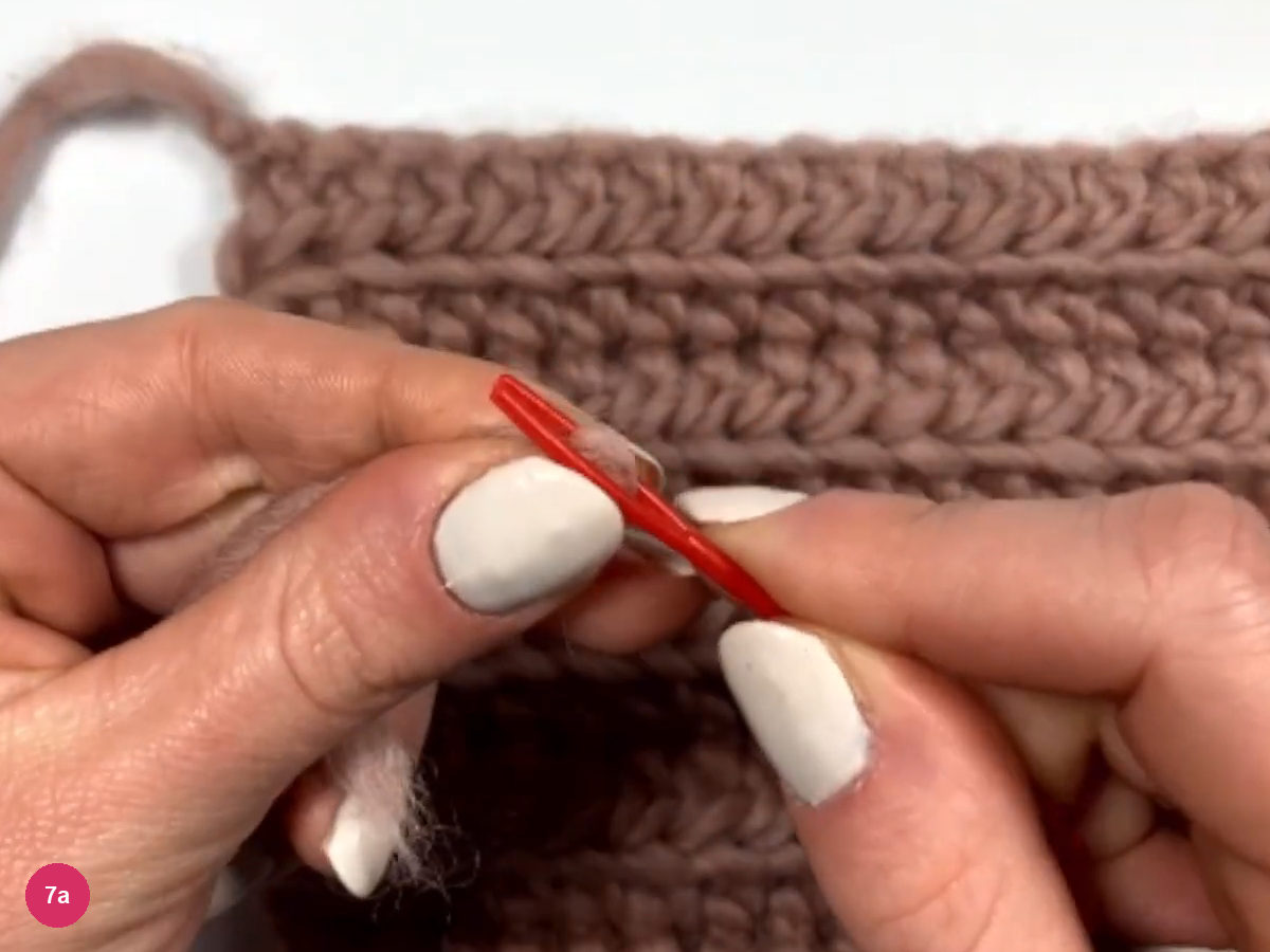 Fingers holding a red tapestry needle near a rose yarn tail with the ribbed panel behind