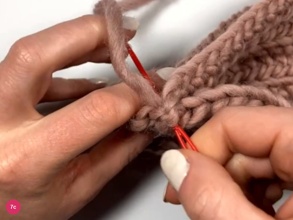 Red needle passing through the seam edge with yarn pulled through, held between both hands