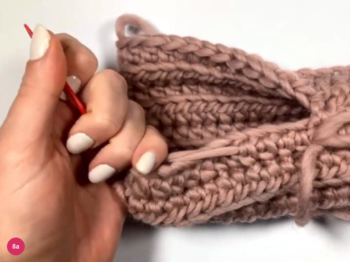 Hand holding the red needle with the other tail threaded, beside the half-sewn sleeve with its open thumb gap