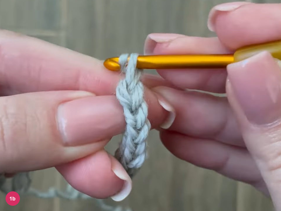 Narrow light grey strip of slip stitches hanging from the hook between fingertips