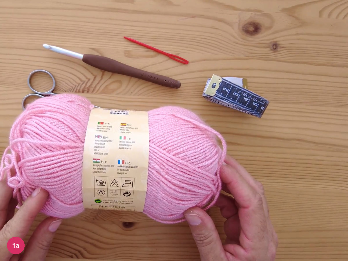 Hands holding a pink yarn ball above a 6 mm crochet hook, a red yarn needle, scissors and a tape measure on a wooden table
