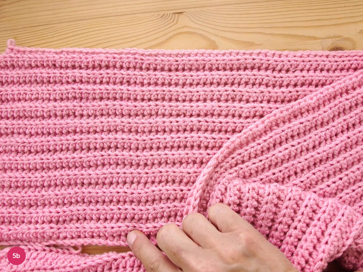 A wide pink ribbed crochet band lying flat on a wooden table, rows clearly countable and one end folded over