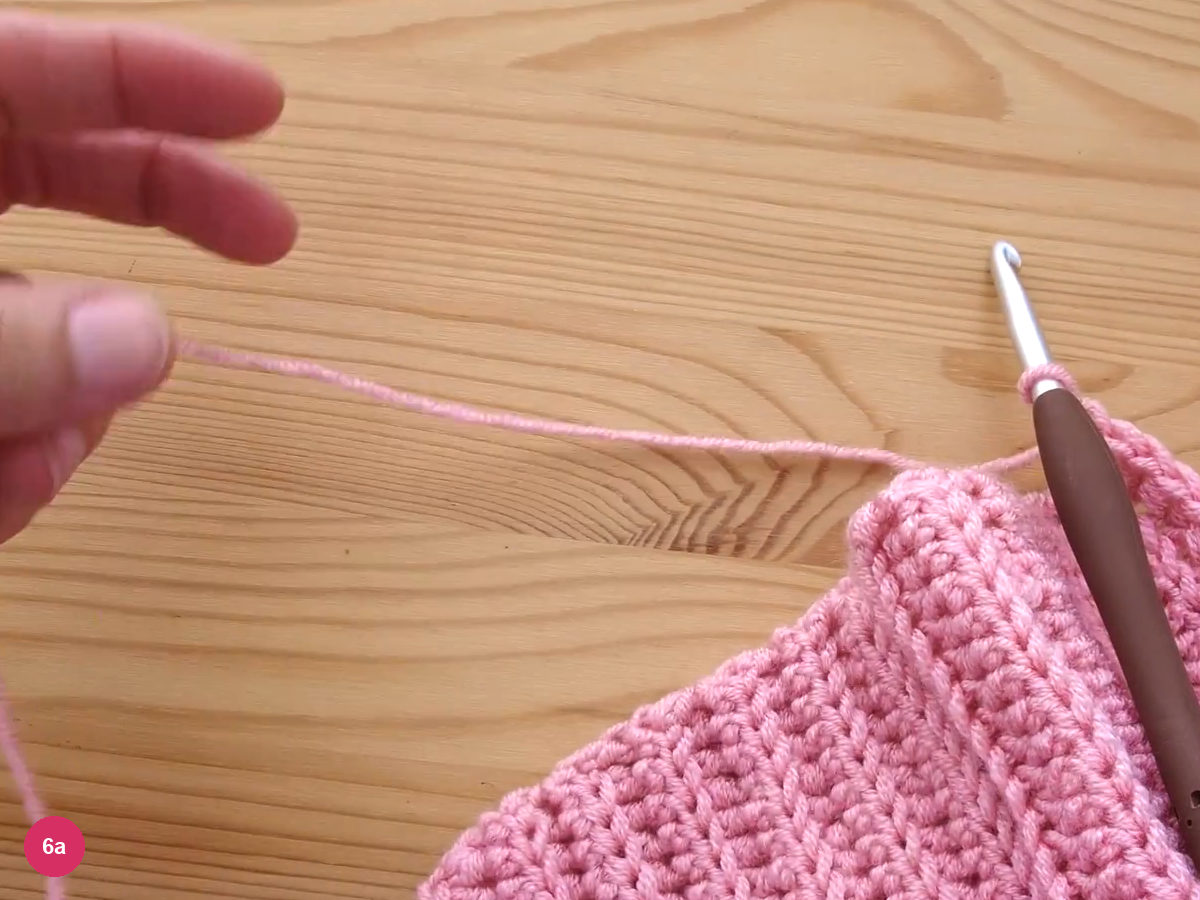 A long pink yarn end pulled out from the last stitch and held up by a hand, the hook lying beside the fabric