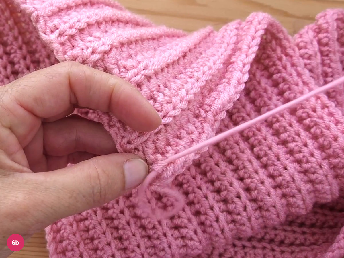 A pink yarn tail pulled out through the stitches at the fabric edge after the needle pass, with a small loop at the end