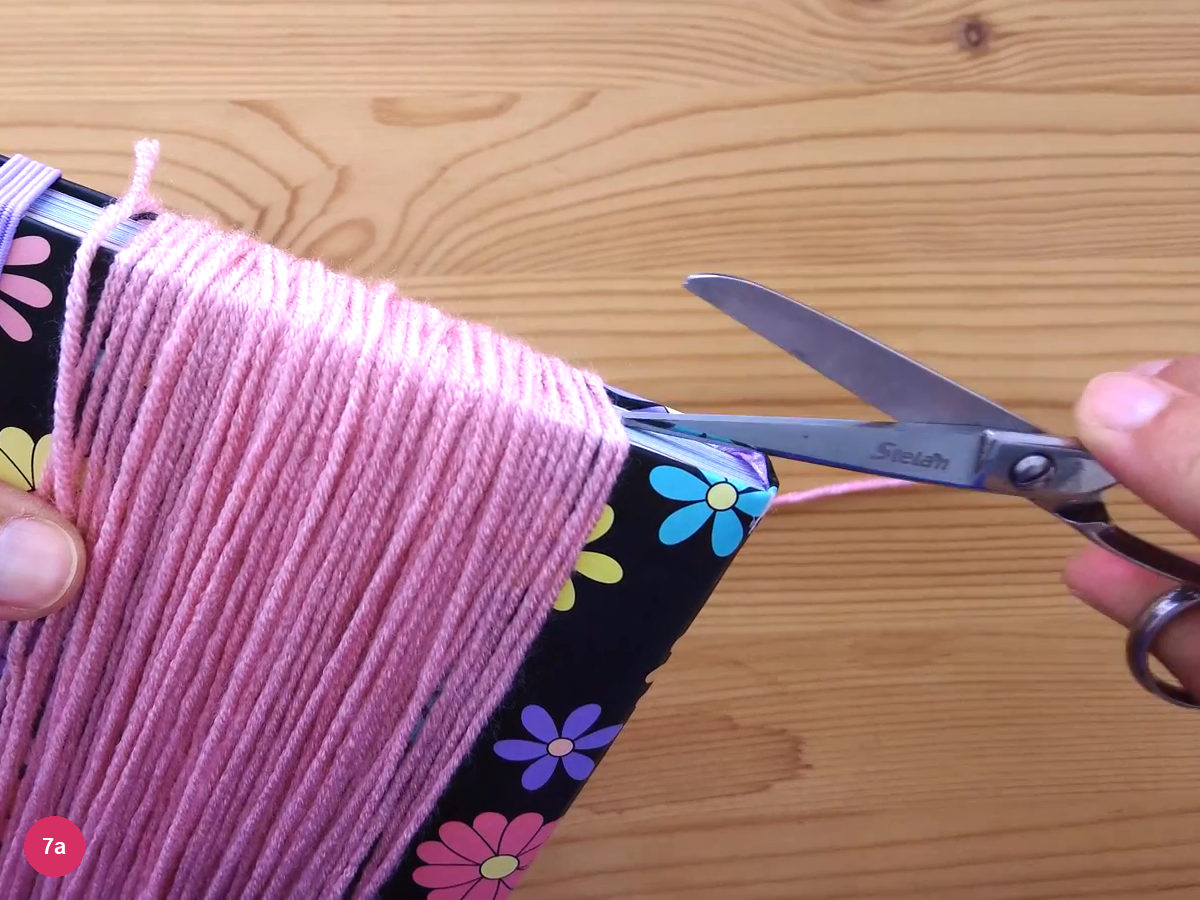 Pink yarn wound around a flat notebook with scissors cutting the strands along one edge