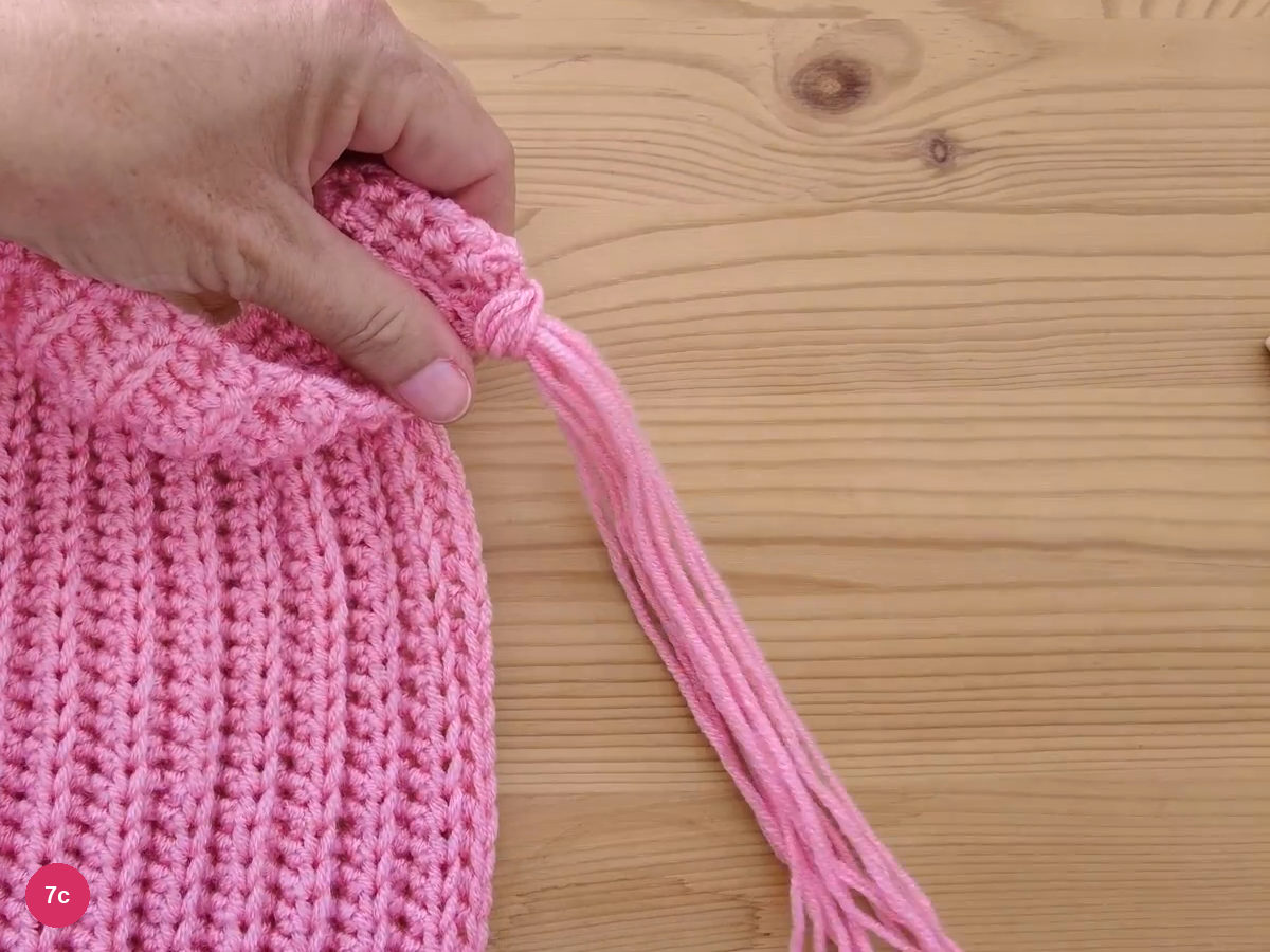 A pink fringe tassel knotted into the scarf edge, its loose ends hanging over the wooden table