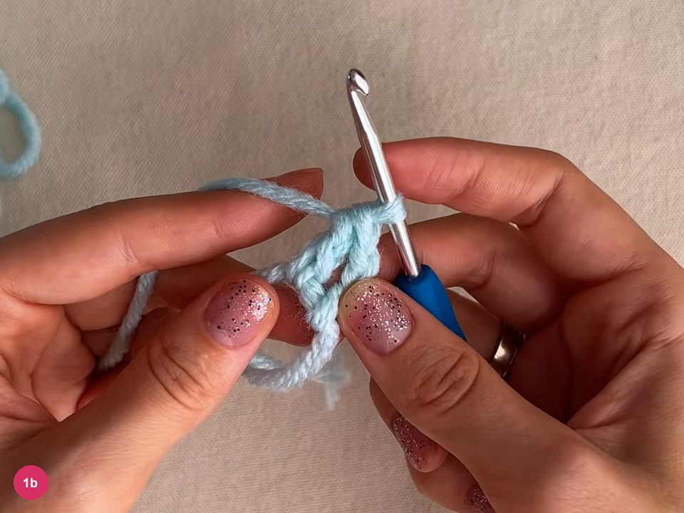 Mint yarn loops and the first stitches of round 1 held between the fingers, with the hook standing at the top