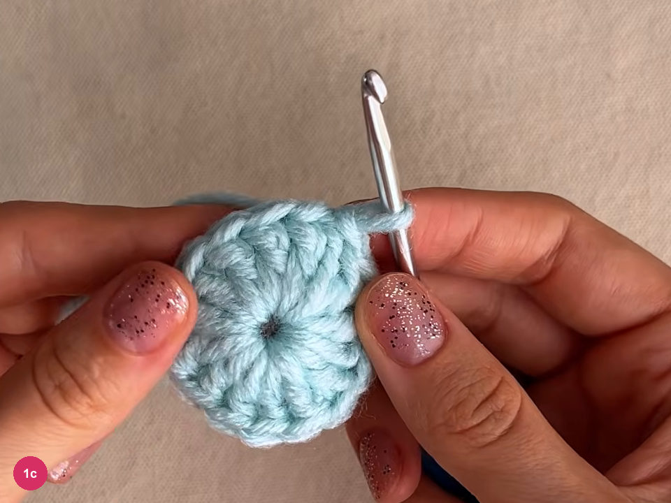 Small closed mint round of dc stitches with a tiny dark centre hole, held by two thumbs, hook at its right edge