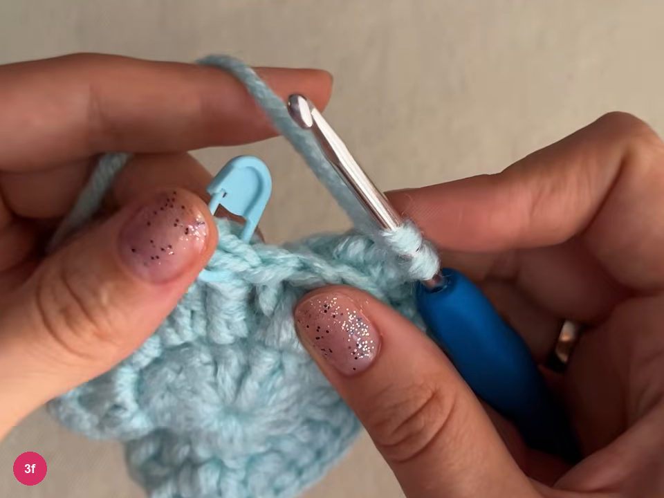 Hook with working yarn at the edge of the mint round, with a blue stitch marker clipped into the work at the upper left