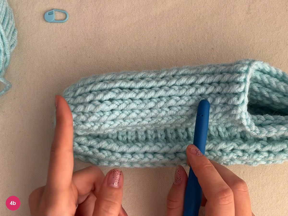 Side view of the mint cup-shaped slipper body with horizontal ribs, a stitch marker at upper left and a hook handle lying on it