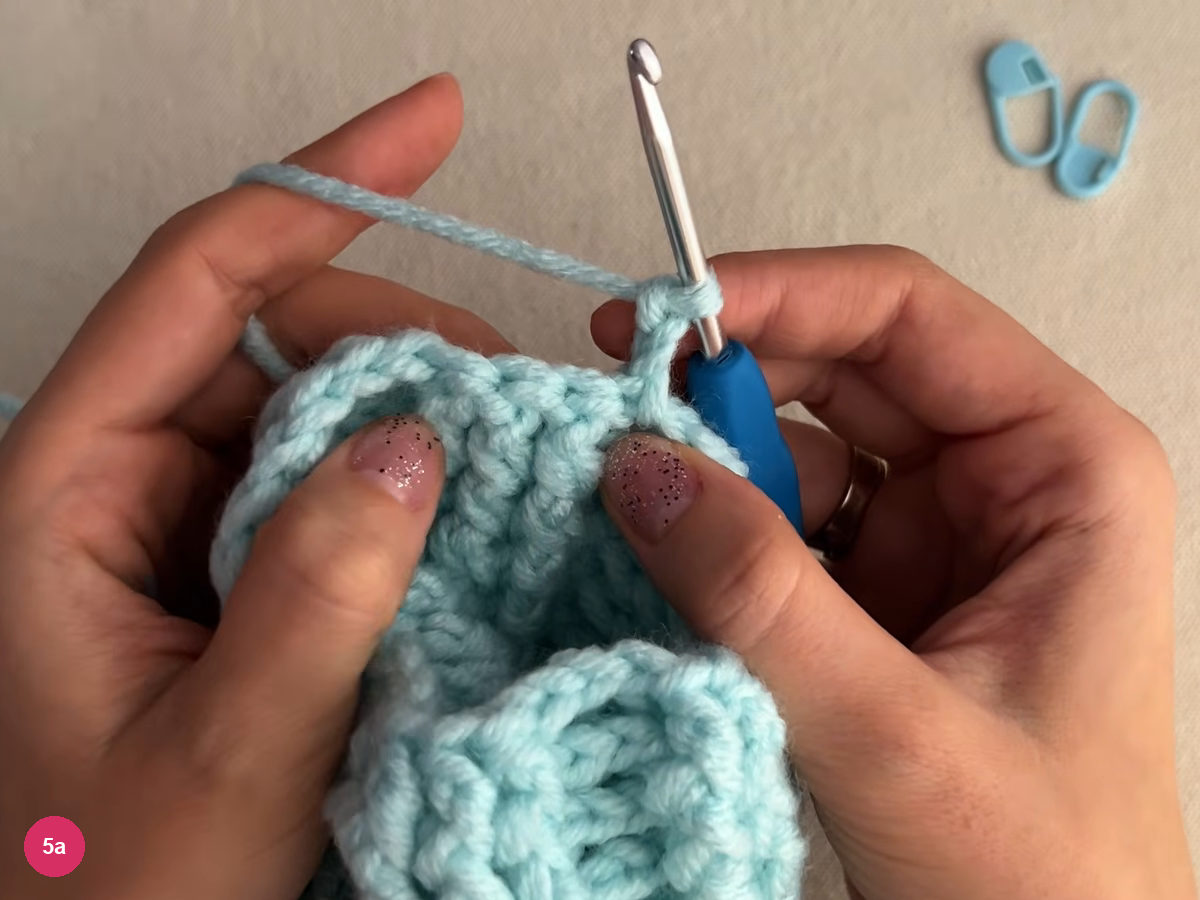 Hook with yarn at the turned edge of the ribbed cup, ribbed rows visible on both sides of the hook and two stitch markers at upper right
